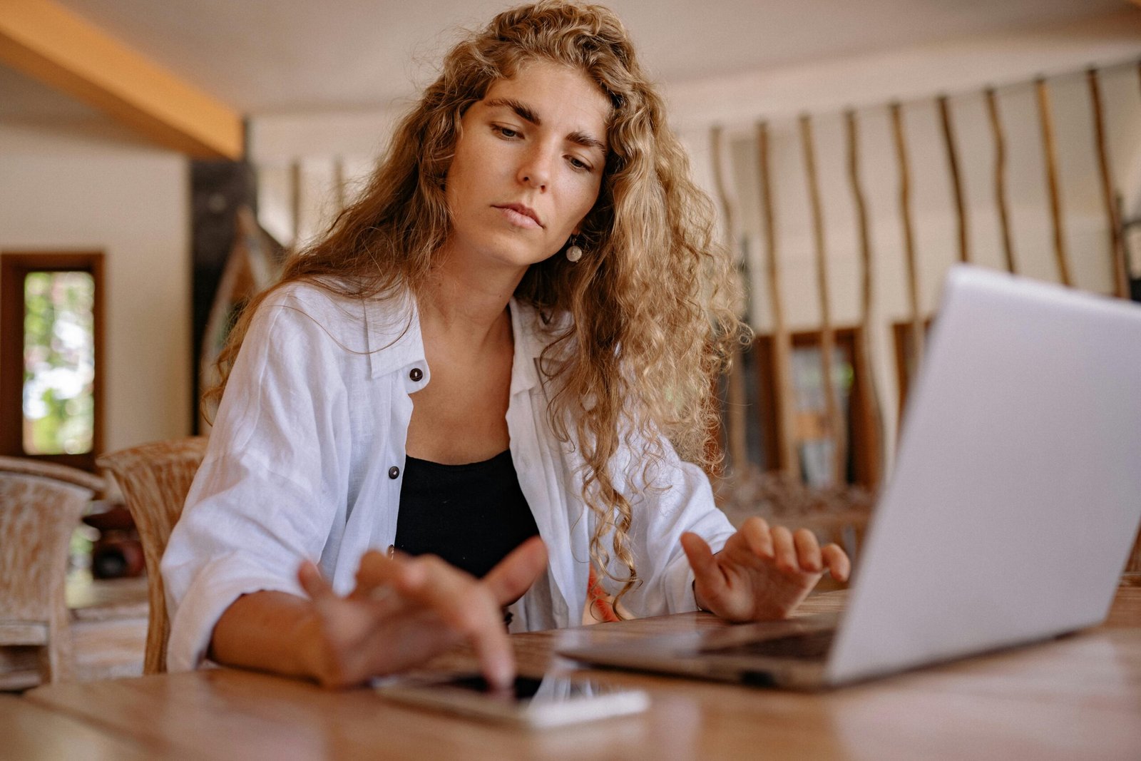 A woman intently works on a laptop while using a smartphone indoors, symbolizing modern remote work.